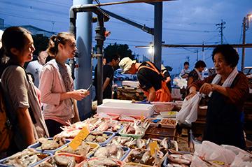 露店に沢山の魚がかごに入れられ並んでいて、お客さんが買い物をしている写真