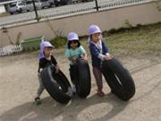 園庭で紫の帽子を被った三人の保育園児がタイヤを転がしている写真