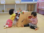 赤ちゃんが2人畳に座っていて、台形の形をしたおもちゃに手を入れて遊んでいる写真