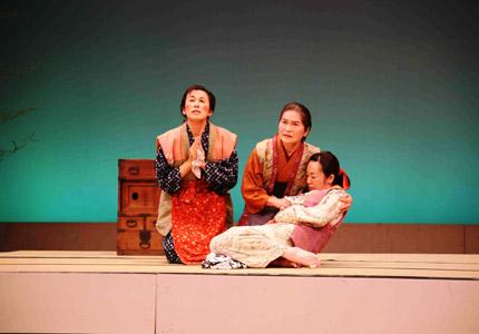 福津民話劇団による舞台で老夫婦が女性を抱きかかえて祈っているシーンの写真