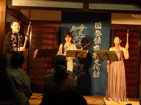 屋敷の中で男性がマイクで話しをし女性2名が木管楽器を持って譜面の前に立っている写真