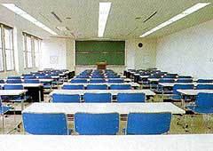 長机に3脚の椅子が並んでいる席が沢山あり、部屋の一番前には黒板と教卓がある研修室内の写真