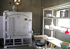 奥には白い陶芸の窯があり、右に道具が置かれた棚がある作業部屋の写真