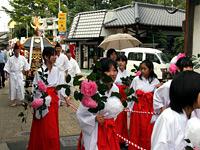 白と赤の着物を着た女性が手にピンク色の花を持っており、後ろには頭に被り物をし、白い着物を着て神輿を担いでいる方々の写真