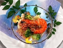 お皿に敷かれたペーパの上に南天の葉が飾られており、その上のガラスの器に調理された海老とブロッコリーやカリフラワーが一緒に盛り付けられている写真